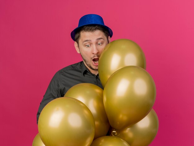 Chico de fiesta joven sorprendido con sombrero azul de pie detrás de globos aislados en rosa