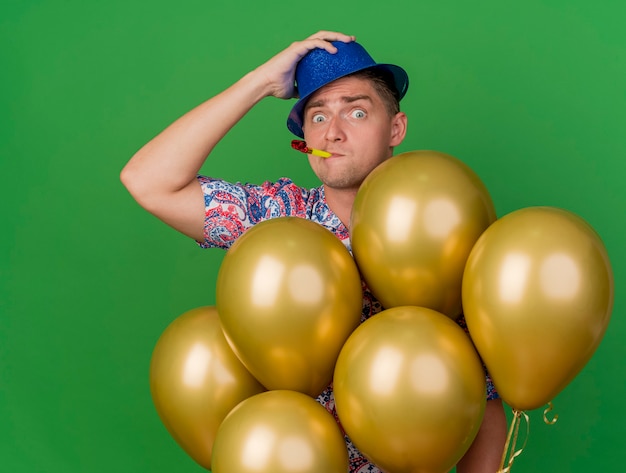 Chico de fiesta joven sorprendido con sombrero azul de pie entre globos poniendo la mano en el sombrero aislado en verde