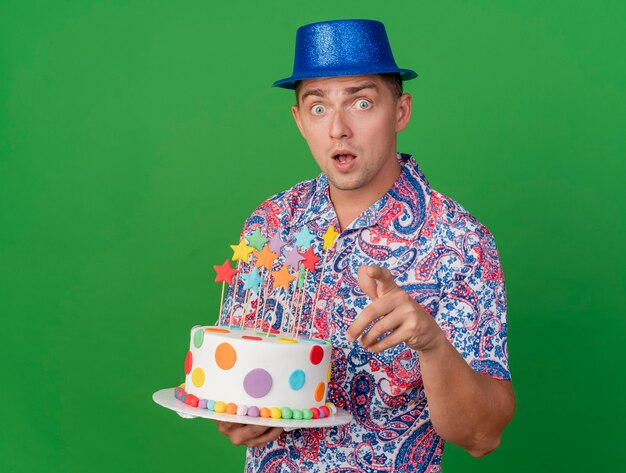 Chico de fiesta joven sorprendido con sombrero azul sosteniendo pastel mostrando gesto aislado en verde