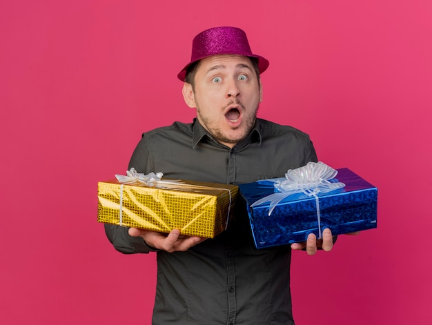 Foto gratuita chico de fiesta joven sorprendido con sombrero rosa con cajas de regalo aisladas en rosa