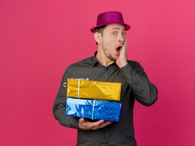 Chico de fiesta joven sorprendido con sombrero rosa sosteniendo cajas de regalo poniendo la mano en la boca aislada en rosa