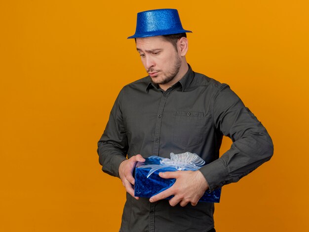 Chico de fiesta joven triste con sombrero azul con caja de regalo aislada en naranja
