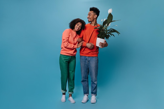 Foto gratuita chico genial y su novia posando con planes sobre fondo azul joven mujer feliz en suéter naranja y hombre moreno en jeans abrazo