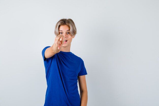 Chico guapo adolescente apuntando hacia adelante con camiseta azul y mirando sorprendido, vista frontal.