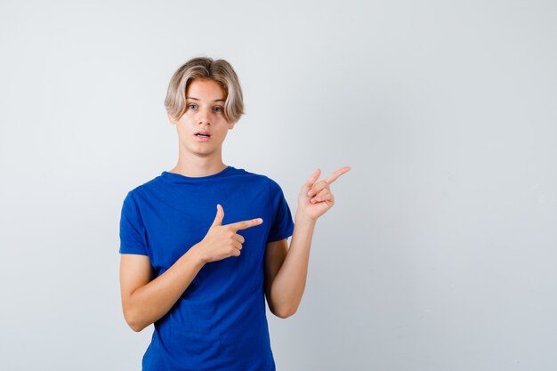 Chico guapo adolescente en camiseta azul apuntando a la esquina superior derecha y mirando perplejo, vista frontal.