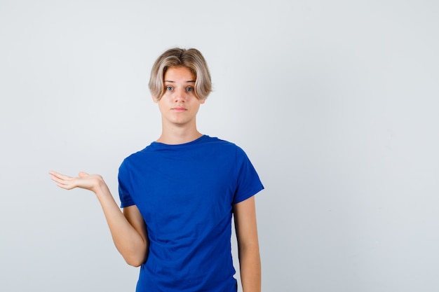 Chico guapo adolescente pretendiendo sostener o mostrar algo en camiseta azul y mirando perplejo, vista frontal.