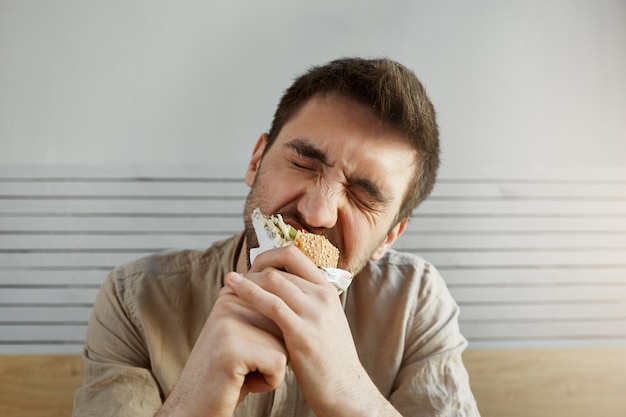 Chico guapo sin afeitar joven con cabello oscuro comiendo sandwich en comida rápida con los ojos cerrados, con expresión feliz y satisfecha.