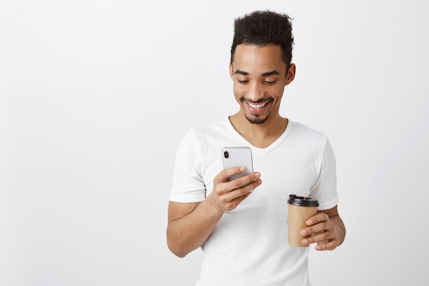 Chico guapo afroamericano en camiseta blanca charlando, enviando mensajes de texto y tomando café, mirando la pantalla del teléfono móvil