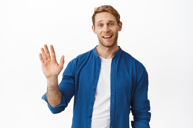 Foto gratuita un chico guapo y amistoso con el pelo rojo te dice hola, te saluda con la mano y sonríe, saluda a un amigo con un gesto de hola, de pie sobre una pared blanca