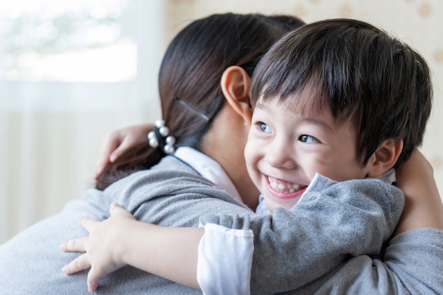 Chico guapo asiático sonriendo felizmente y abrazando con madre en casa, concepto de familia