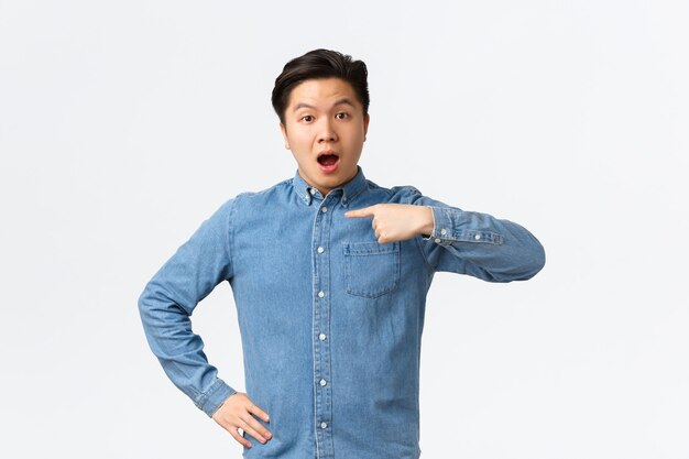 Chico guapo asiático sorprendido y cuestionado con camisa azul, apuntando a sí mismo con cara de curiosidad, siendo mencionado o nombrado, elegido entre la multitud, de pie fondo blanco