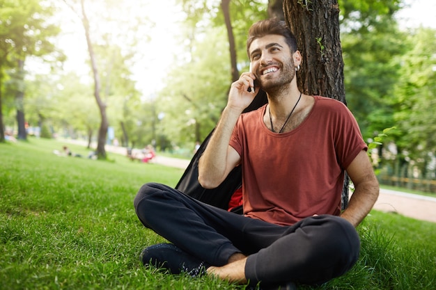 Chico guapo con barba descansando en el parque sobre el césped, hablando por teléfono móvil y sonriendo feliz