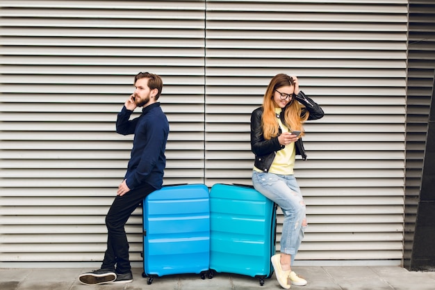 Foto gratuita chico guapo con barba se inclinó sobre la maleta sobre fondo de rayas gris y hablando por teléfono. chica guapa con pelo largo en vasos cerca también se inclinó sobre la maleta y está escribiendo en el teléfono.