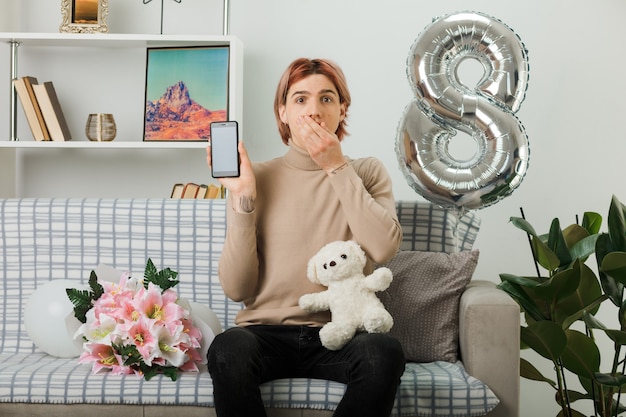 Foto gratuita chico guapo con la boca cubierta de miedo en el día de la mujer feliz con oso de peluche con teléfono sentado en el sofá en la sala de estar