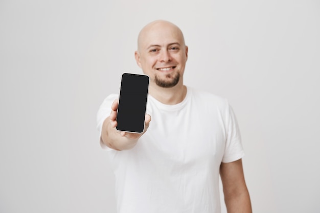 Chico guapo calvo en camiseta blanca que muestra el anuncio de la pantalla del teléfono inteligente sonriendo