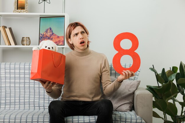 Chico guapo confundido en el día de la mujer feliz sosteniendo el número ocho con bolsa de regalo sentado en el sofá en la sala de estar