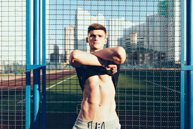 Chico guapo con un cuerpo musculoso desnudándose camiseta negra por la mañana en el estadio. Está mirando a la cámara con confianza.