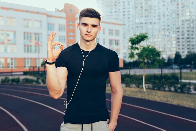 Chico guapo con un cuerpo musculoso posando para la cámara por la mañana en el estadio. Lleva camiseta negra, auriculares. Está mirando a la cámara con confianza.