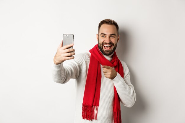 Chico guapo deseando feliz navidad en videollamada, saludando con la mano en el teléfono móvil y sonriendo, de pie en suéter con pañuelo rojo, fondo blanco.
