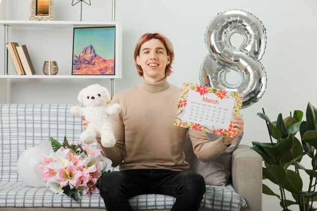 Chico guapo en el día de la mujer feliz con oso de peluche con calendario sentado en el sofá en la sala de estar