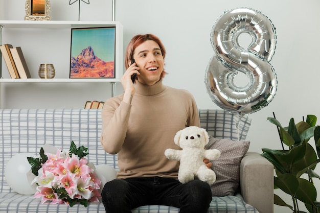 Foto gratuita chico guapo en el día de la mujer feliz con oso de peluche habla por teléfono sentado en el sofá en la sala de estar