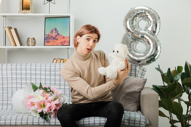 Chico guapo en el día de la mujer feliz con oso de peluche sentado en el sofá en la sala de estar