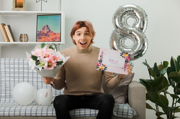 Chico guapo en el día de la mujer feliz con ramo con postal sentado en el sofá en la sala de estar