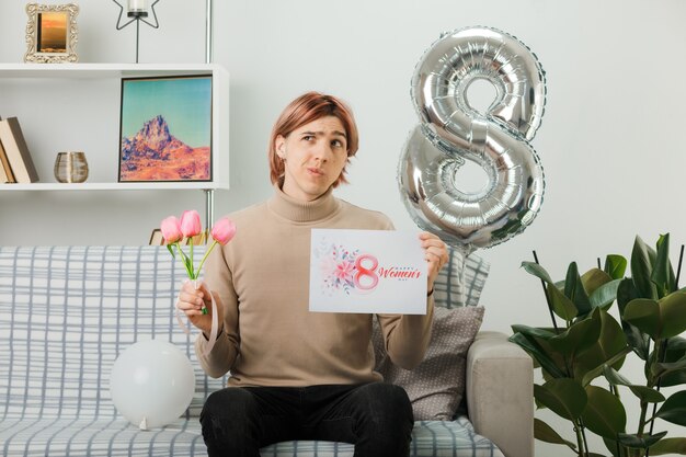 Chico guapo en el día de la mujer feliz sosteniendo flores con postal sentado en el sofá en la sala de estar