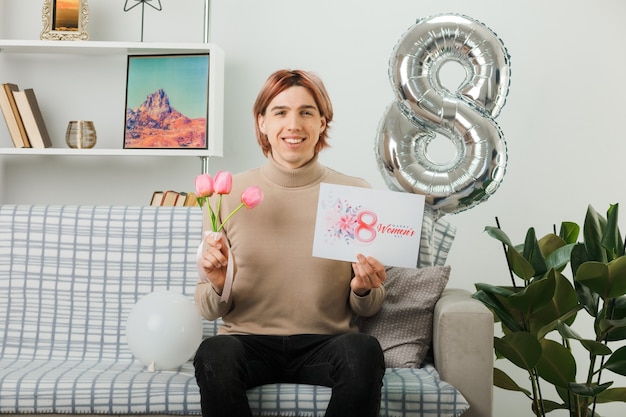 Chico guapo en el día de la mujer feliz sosteniendo flores con postal sentado en el sofá en la sala de estar