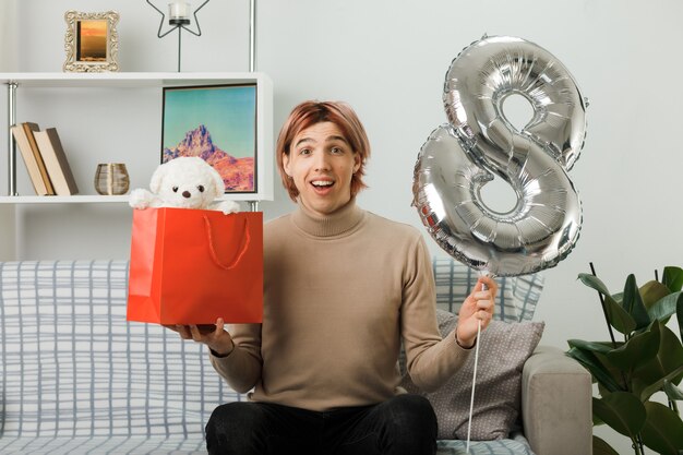 Chico guapo en el día de la mujer feliz sosteniendo el globo número ocho con bolsa de regalo sentado en el sofá en la sala de estar