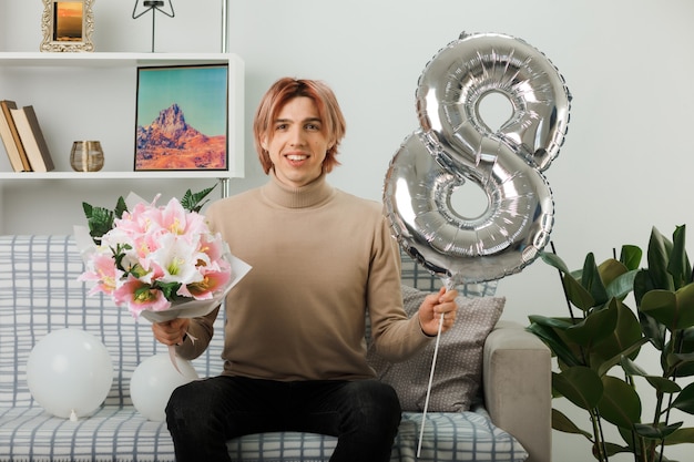 Chico guapo en el día de la mujer feliz sosteniendo el globo número ocho y el ramo sentado en el sofá en la sala de estar