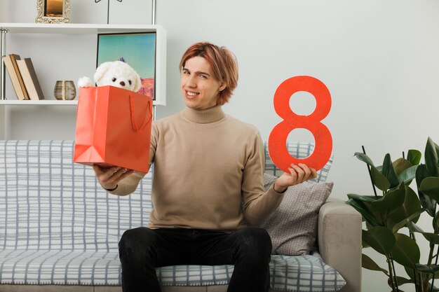 Chico guapo en el día de la mujer feliz sosteniendo el número ocho con bolsa de regalo en la cámara sentado en el sofá en la sala de estar