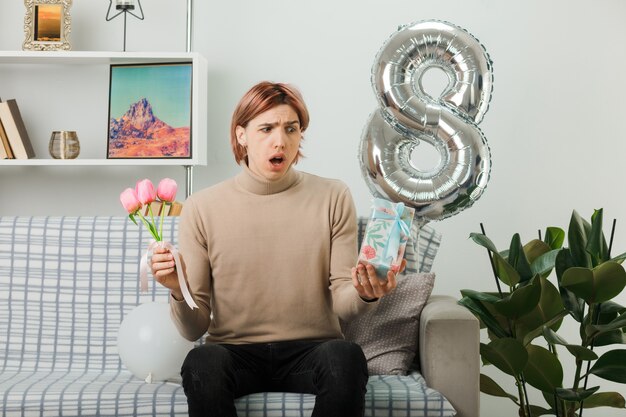 Chico guapo disgustado en el día de la mujer feliz sosteniendo flores con presente sentado en el sofá en la sala de estar