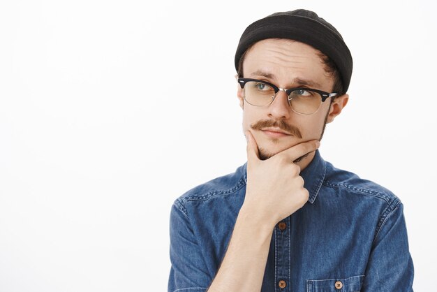 Chico guapo elegante inteligente y creativo pensativo con barba y bigote en gorro negro y gafas mirando a la izquierda con expresión concentrada frotando la barbilla, pensando considerando estar de acuerdo en el trato