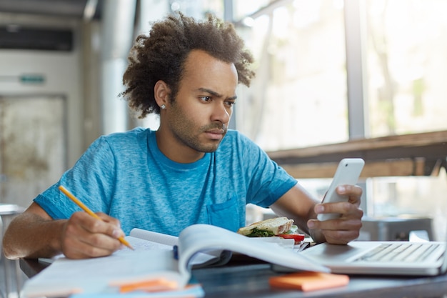 Chico guapo elegante con piel oscura sentado en la cafetería escribiendo algo en su libro de texto con teléfono celular navegando por las noticias a través de las redes sociales en línea y mirando perplejo al descubrir malas noticias