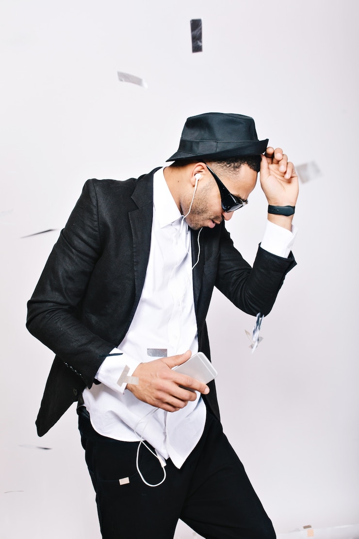 Foto gratuito chico guapo emocionado de moda en traje, sombrero, gafas de sol negras divirtiéndose en oropel. escuchar música a través de headhones, bailar, celebrar la fiesta, genial.