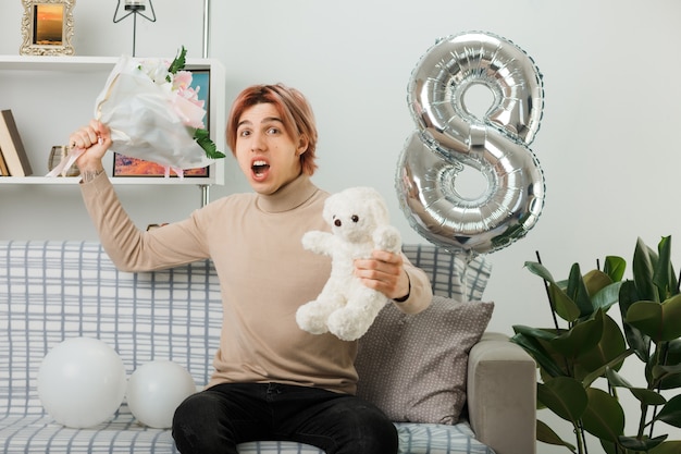 Chico guapo enojado en el día de la mujer feliz con ramo de flores con osito de peluche sentado en el sofá en la sala de estar