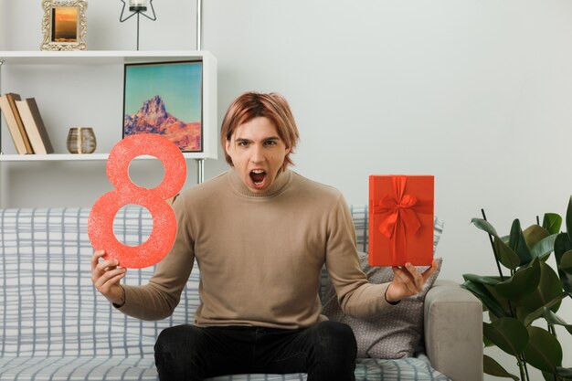 Chico guapo enojado en el día de la mujer feliz sosteniendo el número ocho con presente sentado en el sofá en la sala de estar