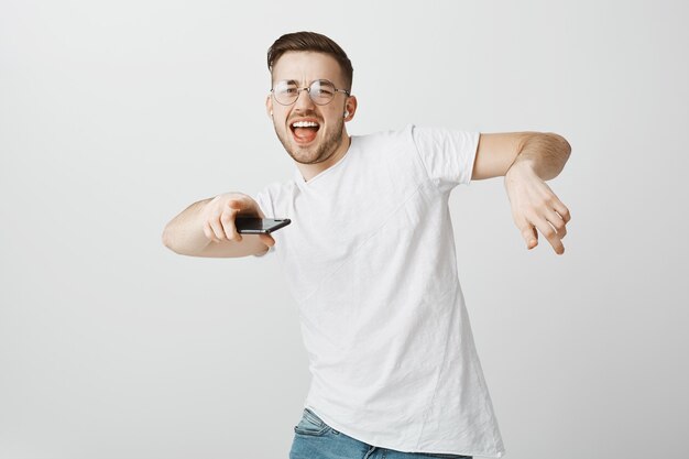 Chico guapo feliz en gafas bailando con música en auriculares inalámbricos con teléfono móvil en la mano