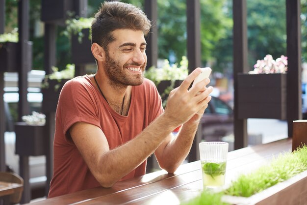 Chico guapo feliz sentado en la cafetería, bebiendo limonada y usando el teléfono móvil, mensaje de texto
