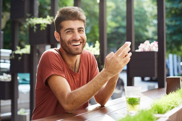 Chico guapo feliz sentado en la cafetería, bebiendo limonada y usando el teléfono móvil, riendo por mensaje de texto