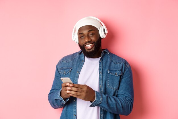 Chico guapo hipster en auriculares sonriendo satisfecho a la cámara, escuchando música en auriculares, usando la aplicación móvil, de pie sobre fondo rosa.