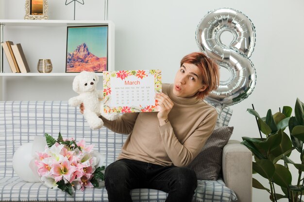 Chico guapo impresionado en el día de la mujer feliz con oso de peluche con calendario sentado en el sofá en la sala de estar