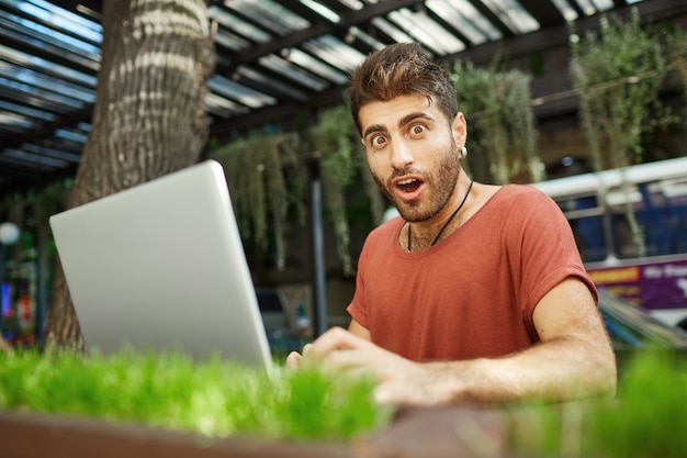 Chico guapo impresionado trabajando al aire libre, autónomo con laptop sentarse en el parque y mirar maravillado