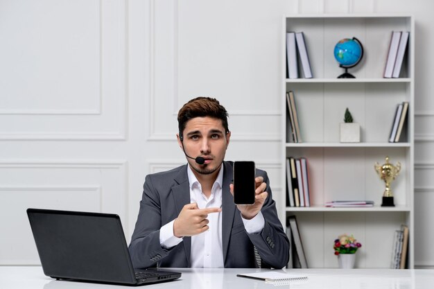 Chico guapo inteligente de servicio al cliente con una computadora portátil en un blazer de oficina gris sosteniendo un teléfono celular