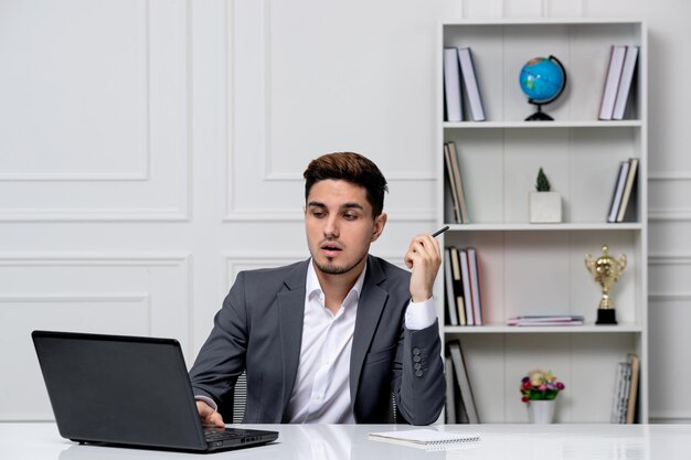 Chico guapo inteligente de servicio al cliente con laptop en blazer gris mirando la pantalla con lápiz