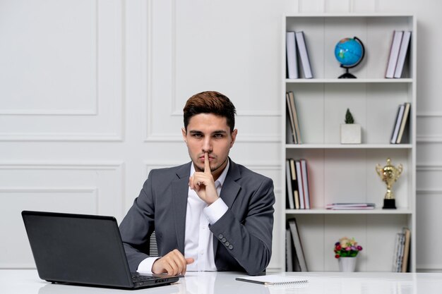 Chico guapo inteligente de servicio al cliente con laptop en blazer de oficina gris que muestra gesto de silencio