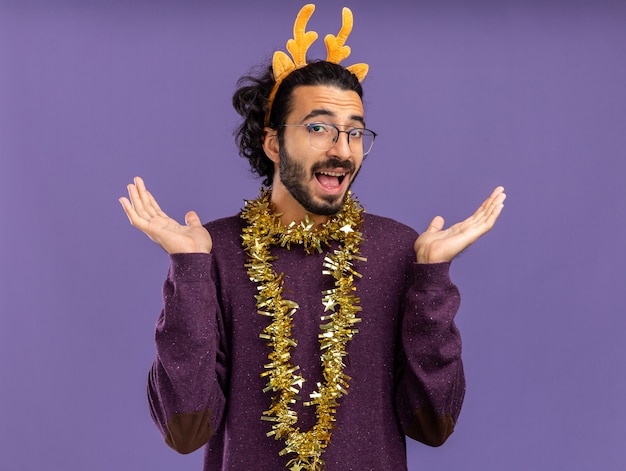 Chico guapo joven alegre con aro de pelo de navidad con guirnalda en el cuello extendiendo las manos aisladas en la pared azul