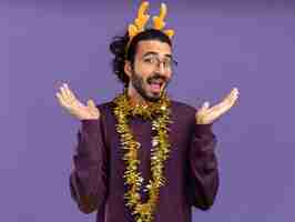 Foto gratuita chico guapo joven alegre con aro de pelo de navidad con guirnalda en el cuello extendiendo las manos aisladas sobre fondo azul.