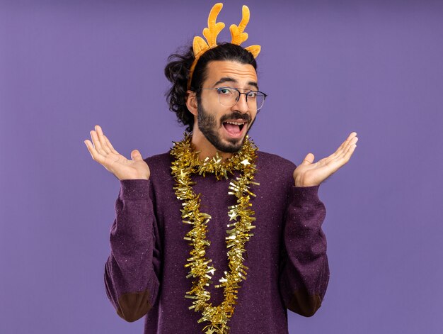 Chico guapo joven alegre con aro de pelo de Navidad con guirnalda en el cuello extendiendo las manos aisladas sobre fondo azul.